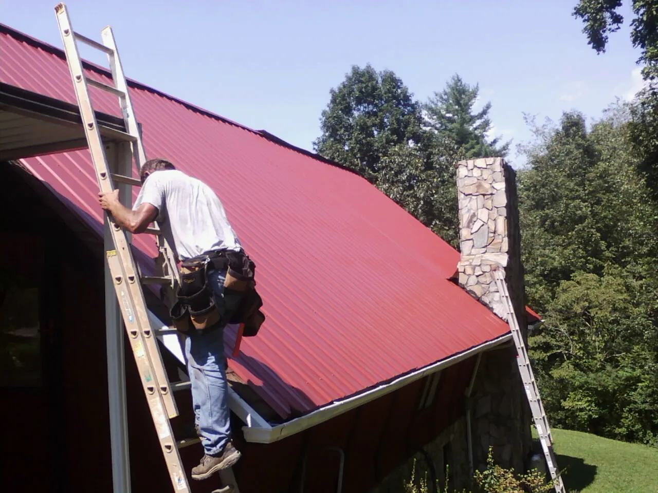 Roofer on a ladder beside a red metal roof and stone chimney — Soffit & Fascia Repair by Roofing Brothers in Purcellville